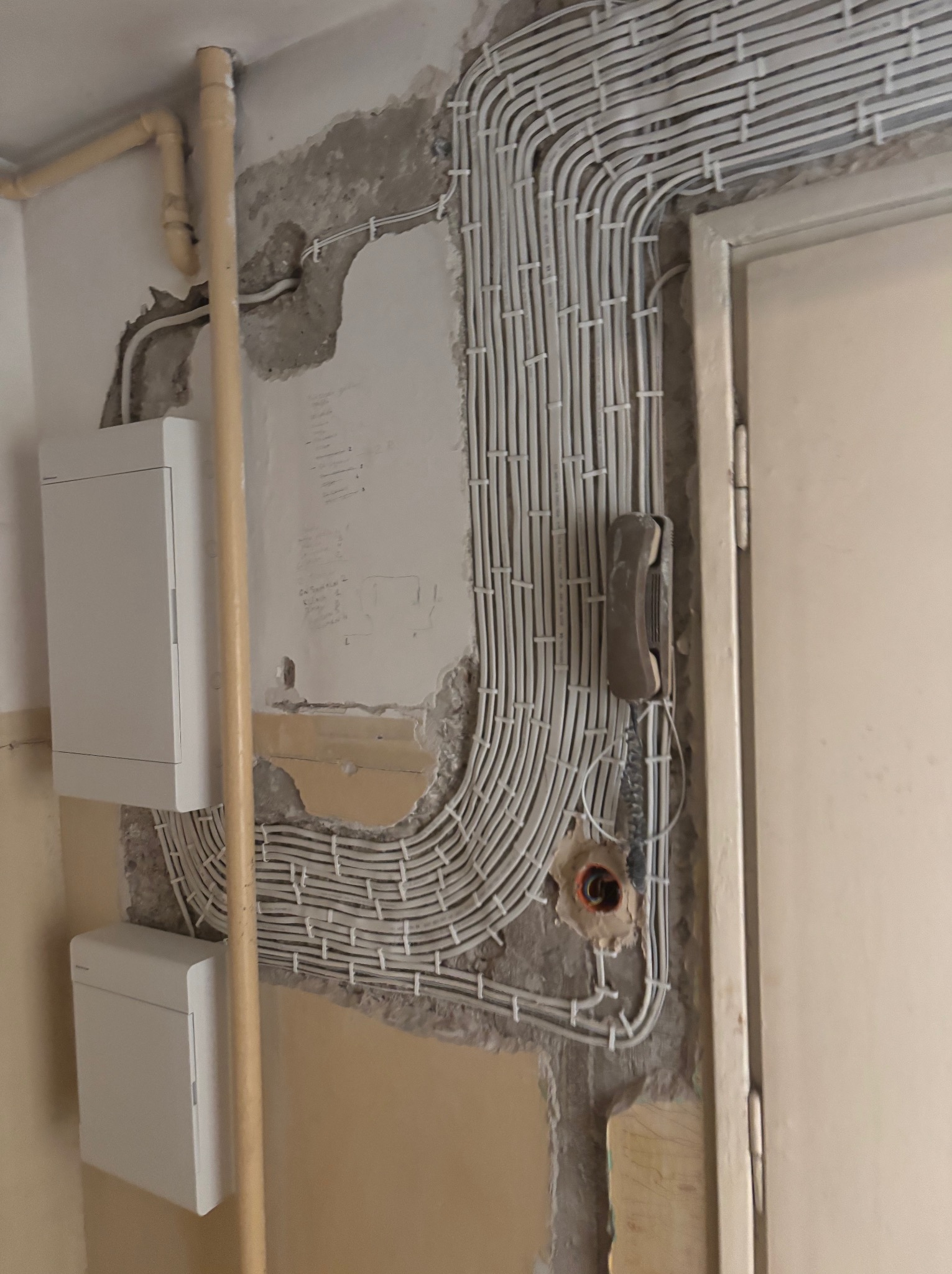 Białe kable elektryczne ułożone równolegle na ścianie w trakcie remontu, obok skrzynka elektryczna i stary domofon. Widoczne ślady po kuciu w ścianie.