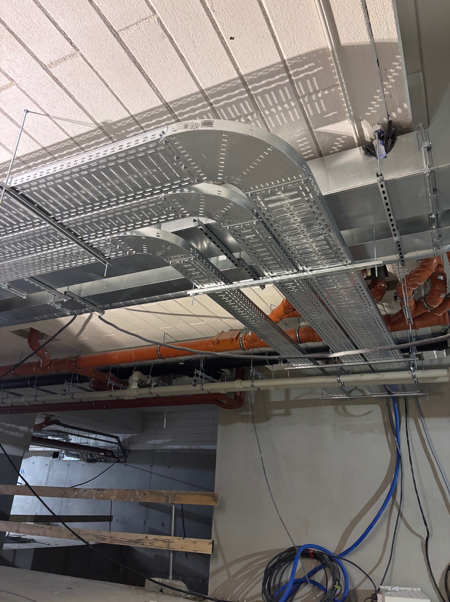 Sufit z zamontowanymi metalowymi korytkami kablowymi i rurami instalacyjnymi w odcieniach pomarańczy i szarości. Widoczne przewody elektryczne w kolorze niebieskim.