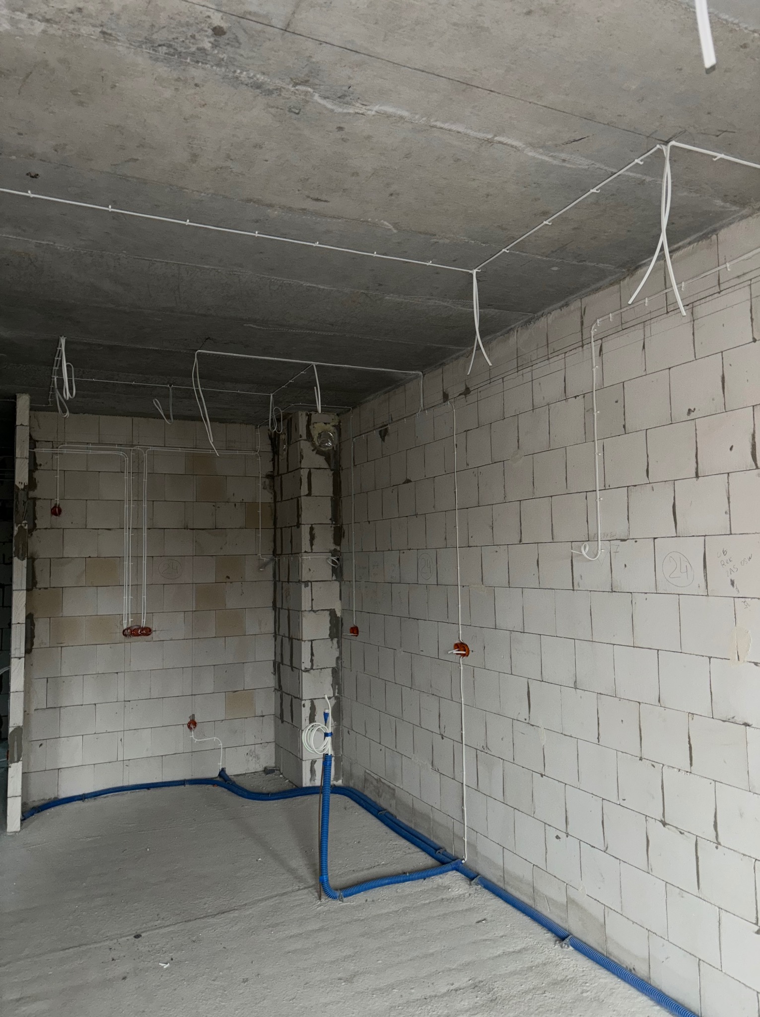 Surowy pokój z instalacją elektryczną: białe kable na suficie i ścianach z czerwonymi puszkami, niebieskie rury na podłodze. Widok na wnętrze w trakcie prac.