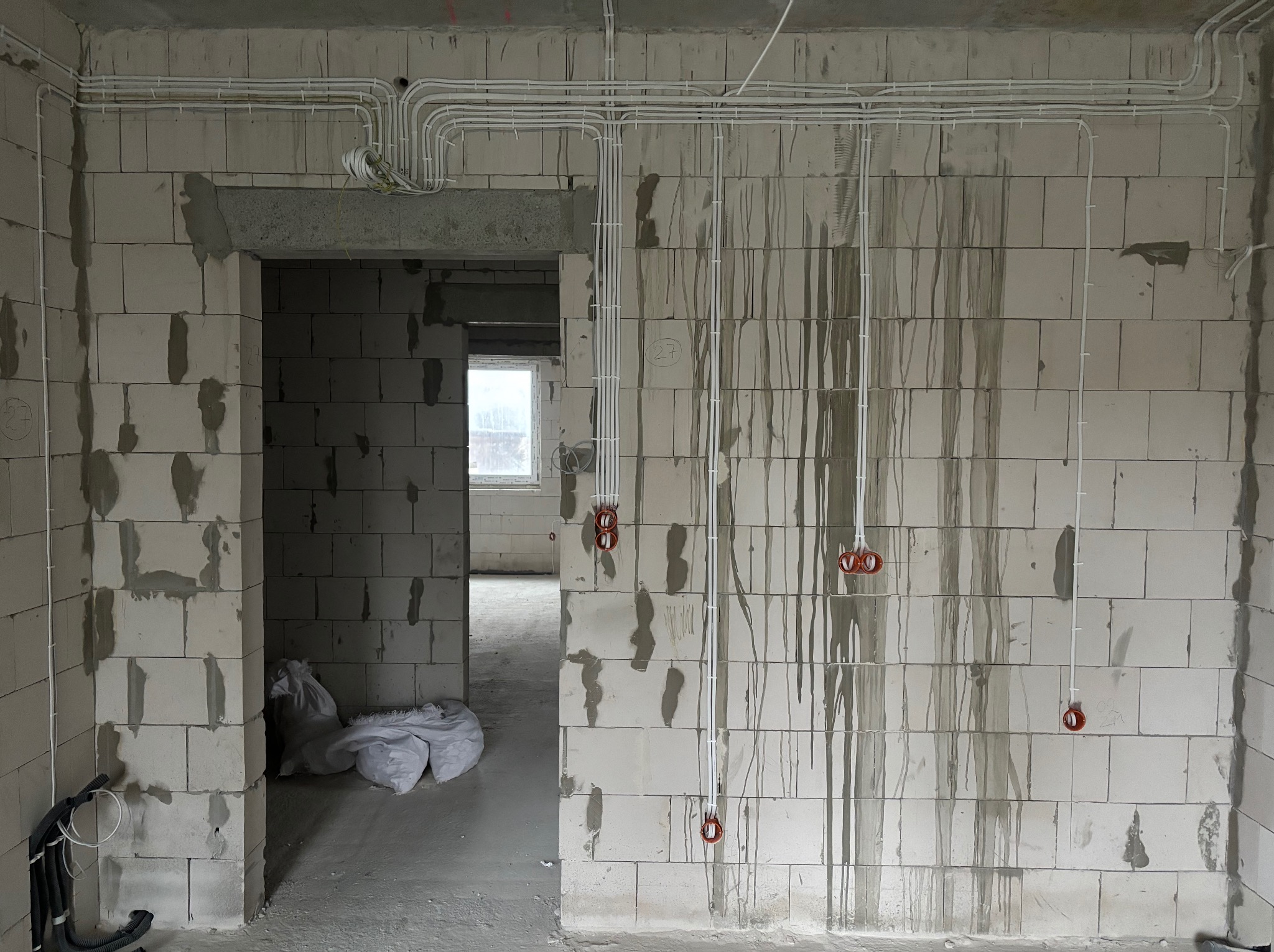 Białe przewody elektryczne ułożone na surowej ścianie z betonu komórkowego wewnątrz budynku w trakcie budowy. Widoczne puszki instalacyjne.