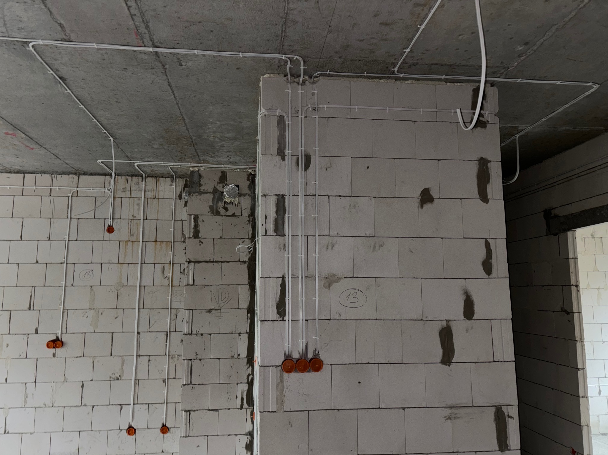 Surowy strop i ściany z betonu komórkowego z widoczną, rozprowadzoną instalacją elektryczną w białych rurkach i pomarańczowymi puszkami. W tle widoczne kolejne pomieszczenie.