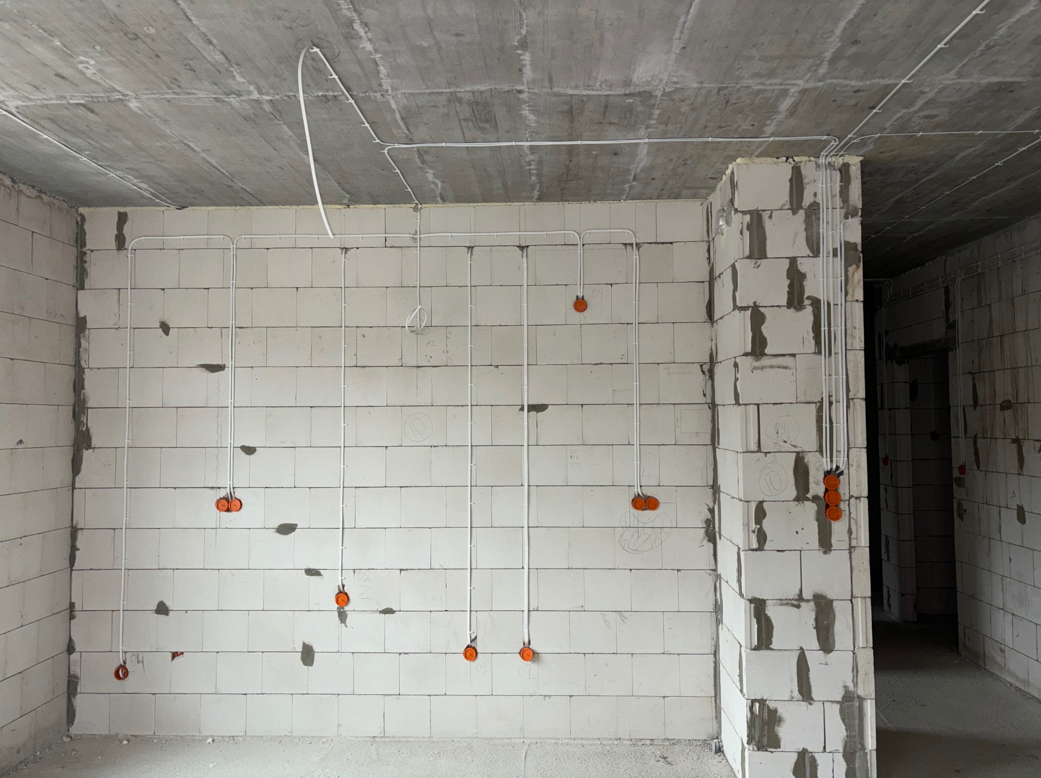 Surowy stan: białe rurki instalacji elektrycznej prowadzone po ścianach z betonu komórkowego, pomarańczowe puszki pod gniazdka i włączniki. Widok wnętrza w trakcie prac.