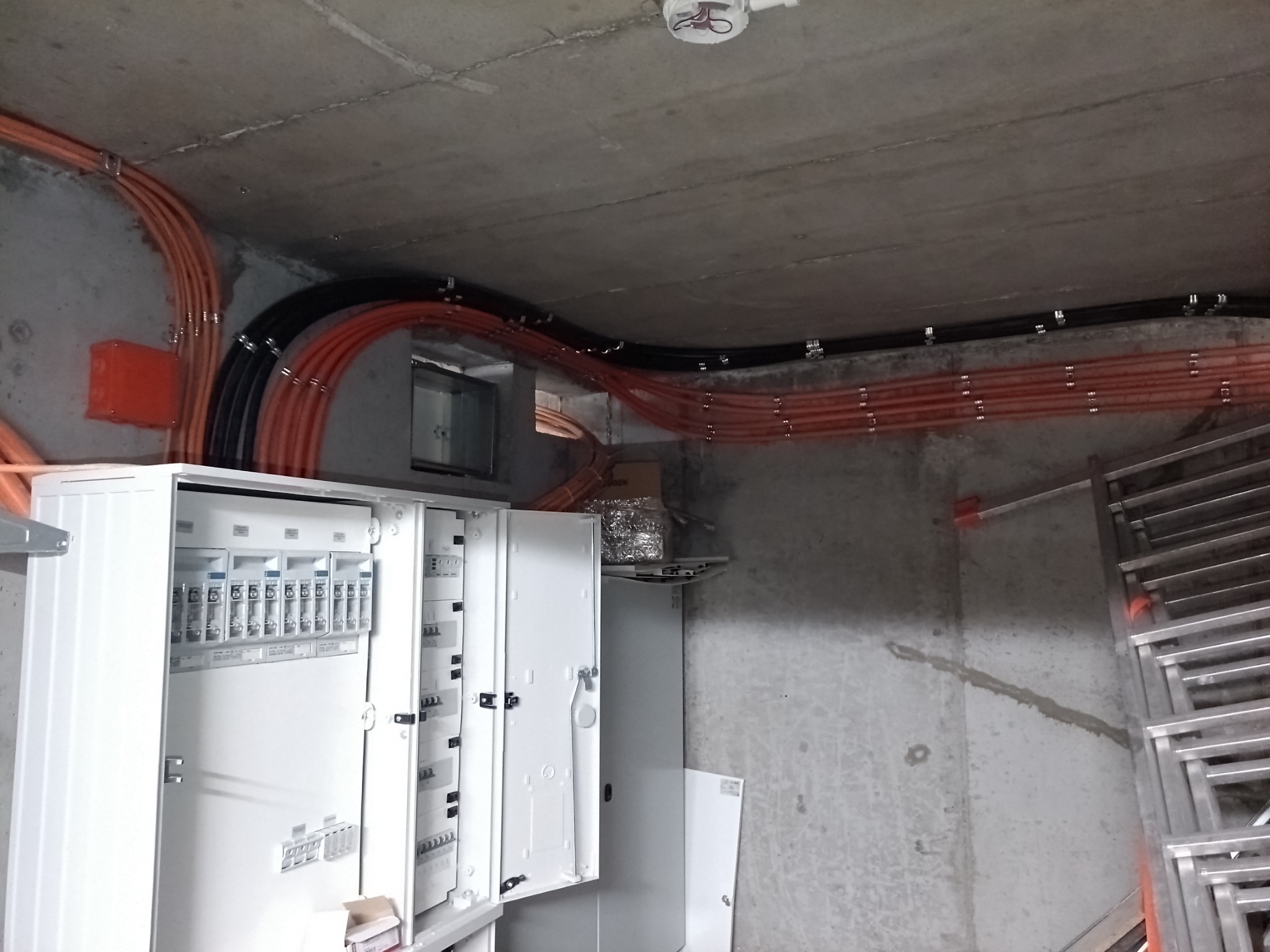 Rozdzielnia elektryczna z widocznymi przewodami w peszlach, zamocowanymi do sufitu. Widoczne zabezpieczenia i aparatura wewnątrz rozdzielnicy.