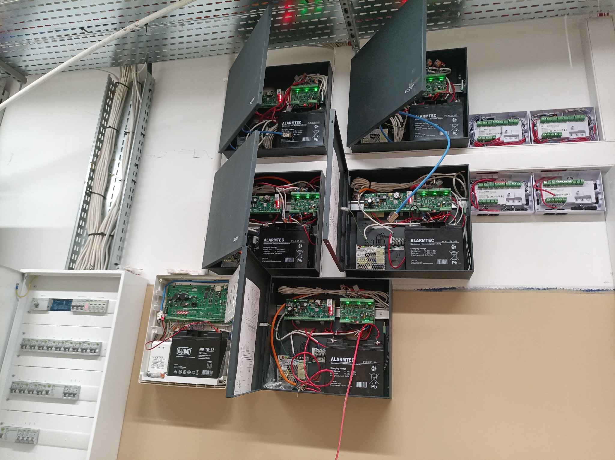 Rozbudowana instalacja alarmowa z otwartymi obudowami, widoczne płyty główne, akumulatory i okablowanie. Dodatkowo skrzynka z bezpiecznikami i kable w korytkach.