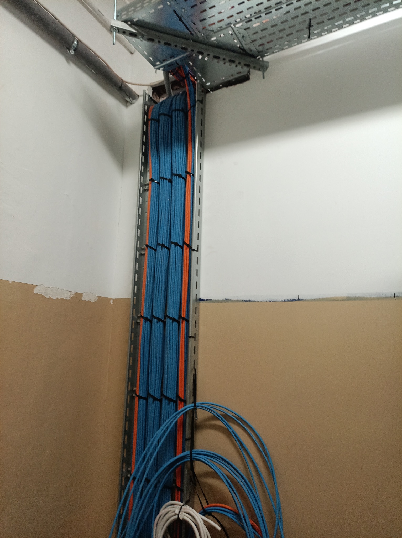 Uporządkowane kable w korycie kablowym, przewody w kolorze niebieskim i pomarańczowym, mocowane czarnymi opaskami. Widoczne fragmenty instalacji pod sufitem.