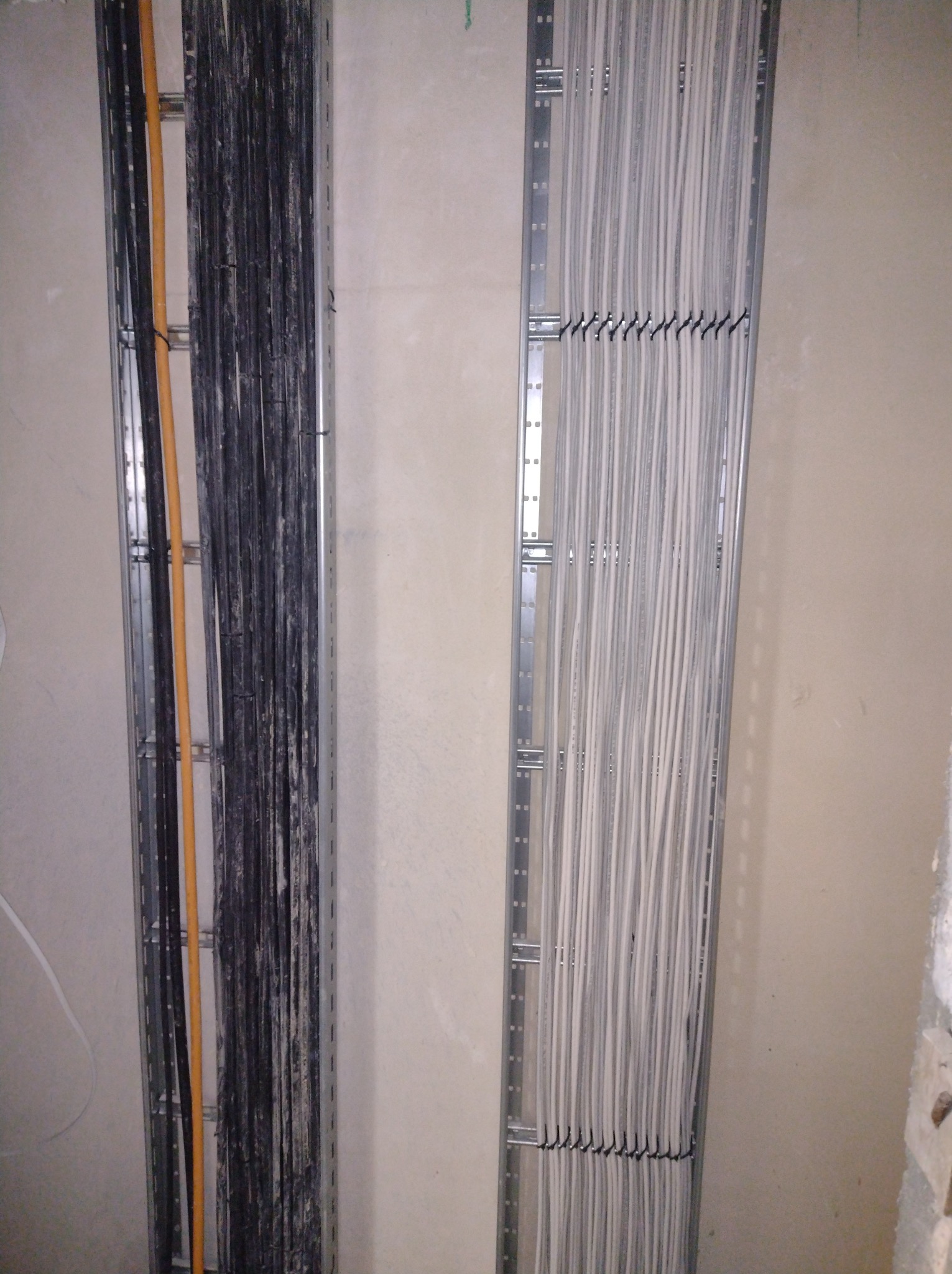 Dwa koryta kablowe zamocowane na ścianie. W jednym korycie ułożone kable w kolorze białym, w drugim czarne i pomarańczowe. Widok z bliska na instalację elektryczną.