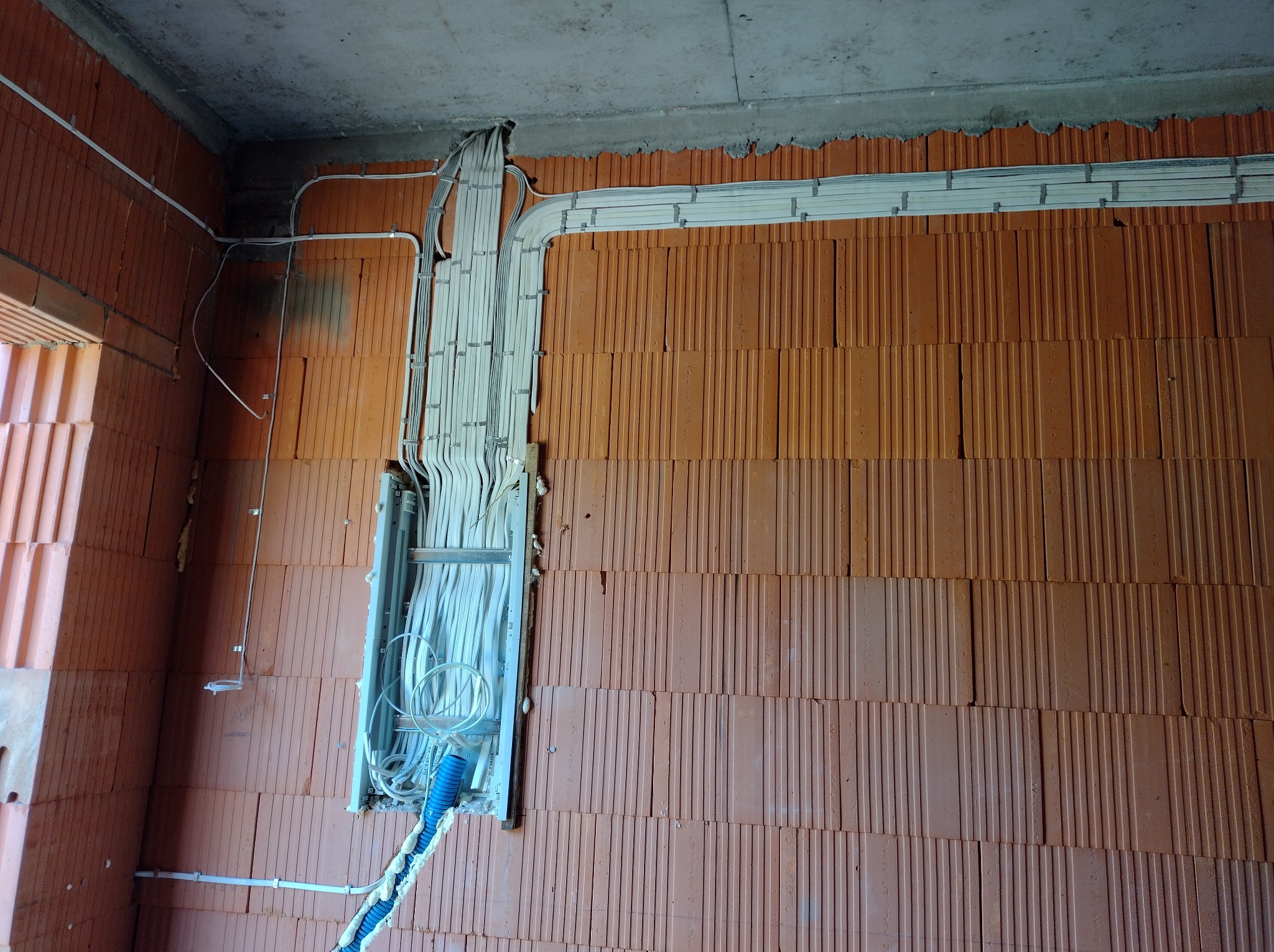 Instalacja elektryczna w trakcie montażu w nowym budynku z cegły. Widoczne kable w białych rurkach przymocowane do ściany i sufitu. Niebieska rura karbowana.