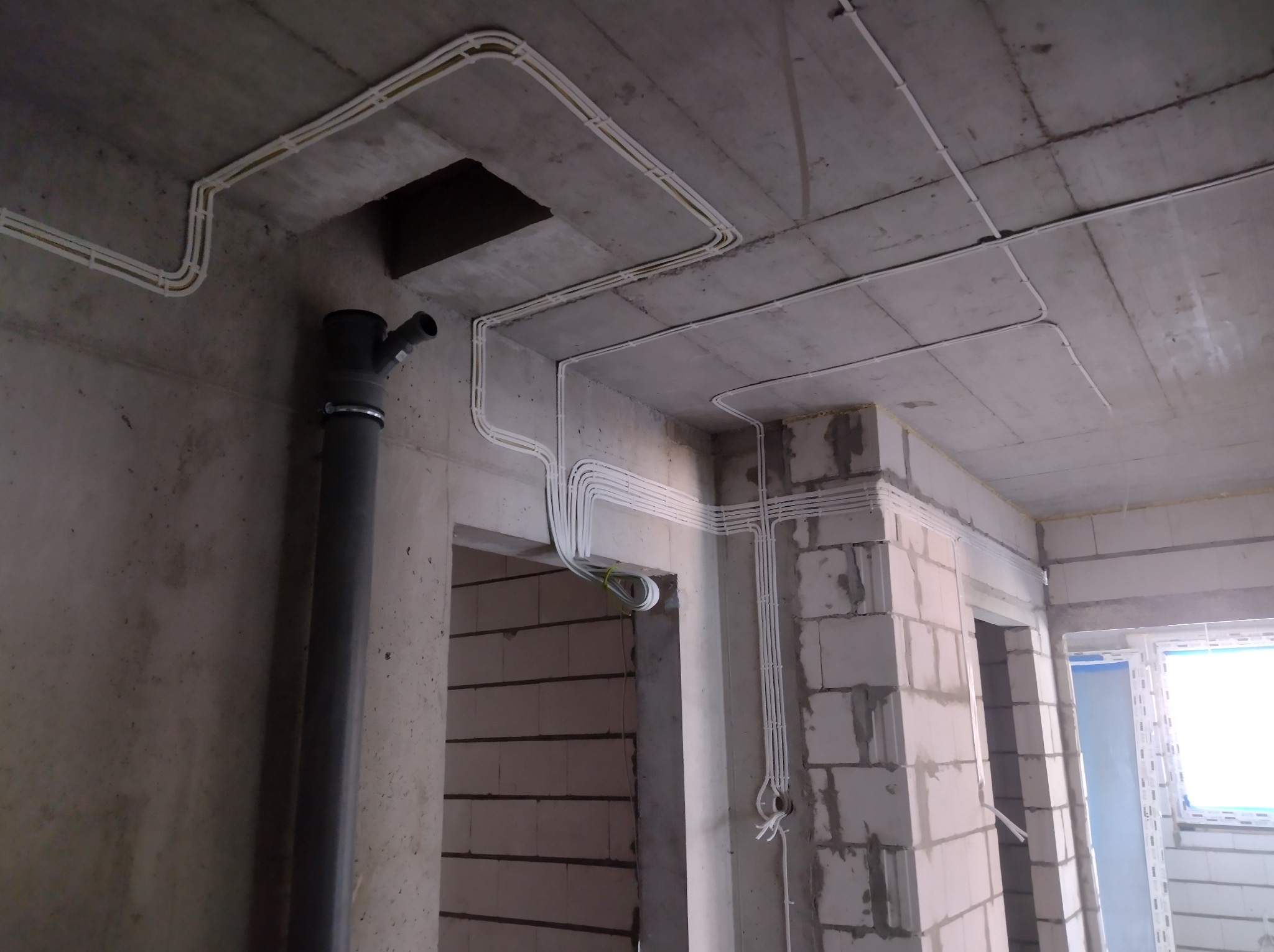 Białe kable elektryczne ułożone w korytkach na surowym, betonowym suficie i ścianach w trakcie budowy. Widoczny pion kanalizacyjny i otwory drzwiowe.