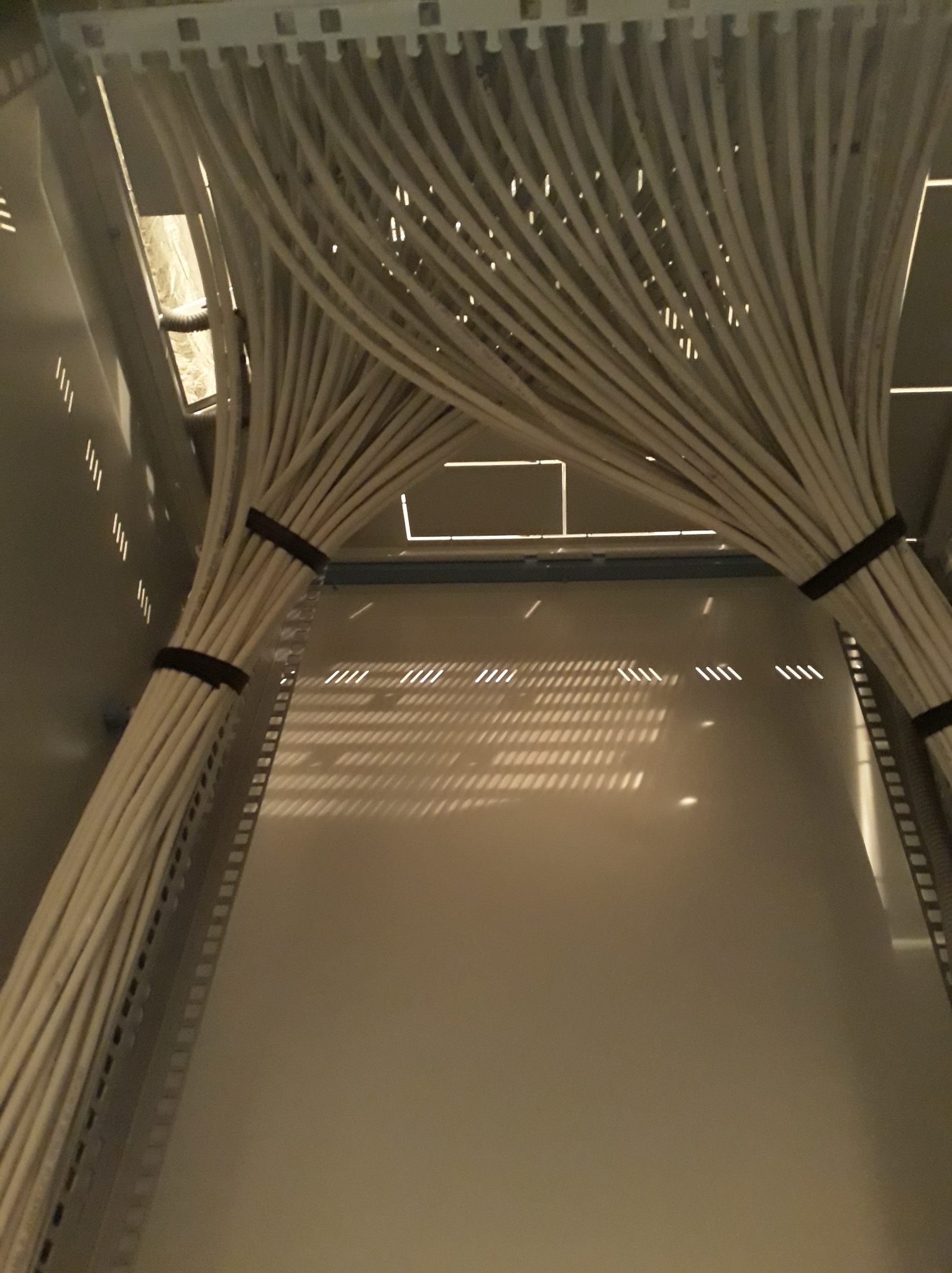 Uporządkowane kable w szafie serwerowej, zebrane w wiązki i przymocowane do konstrukcji. Widoczne perforacje w obudowie szafy i oświetlenie w tle.