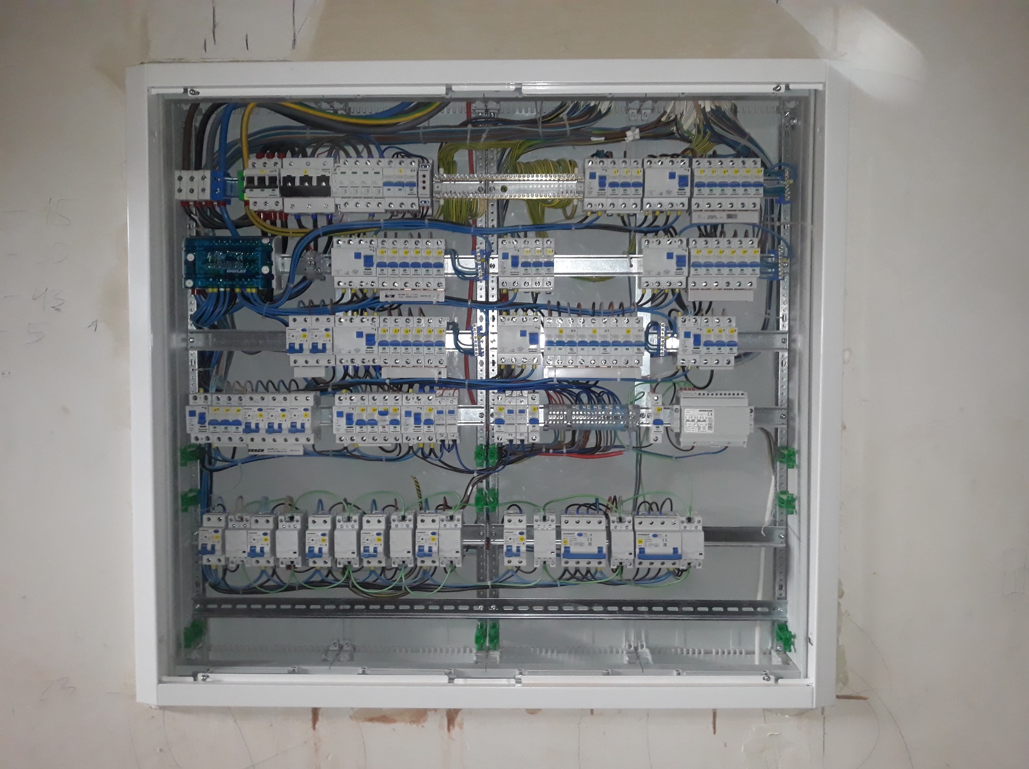 Rozdzielnica elektryczna w białej obudowie, widoczne liczne bezpieczniki, kable i złącza. Montaż w surowym wnętrzu, ściana z widocznymi śladami po pracach budowlanych.