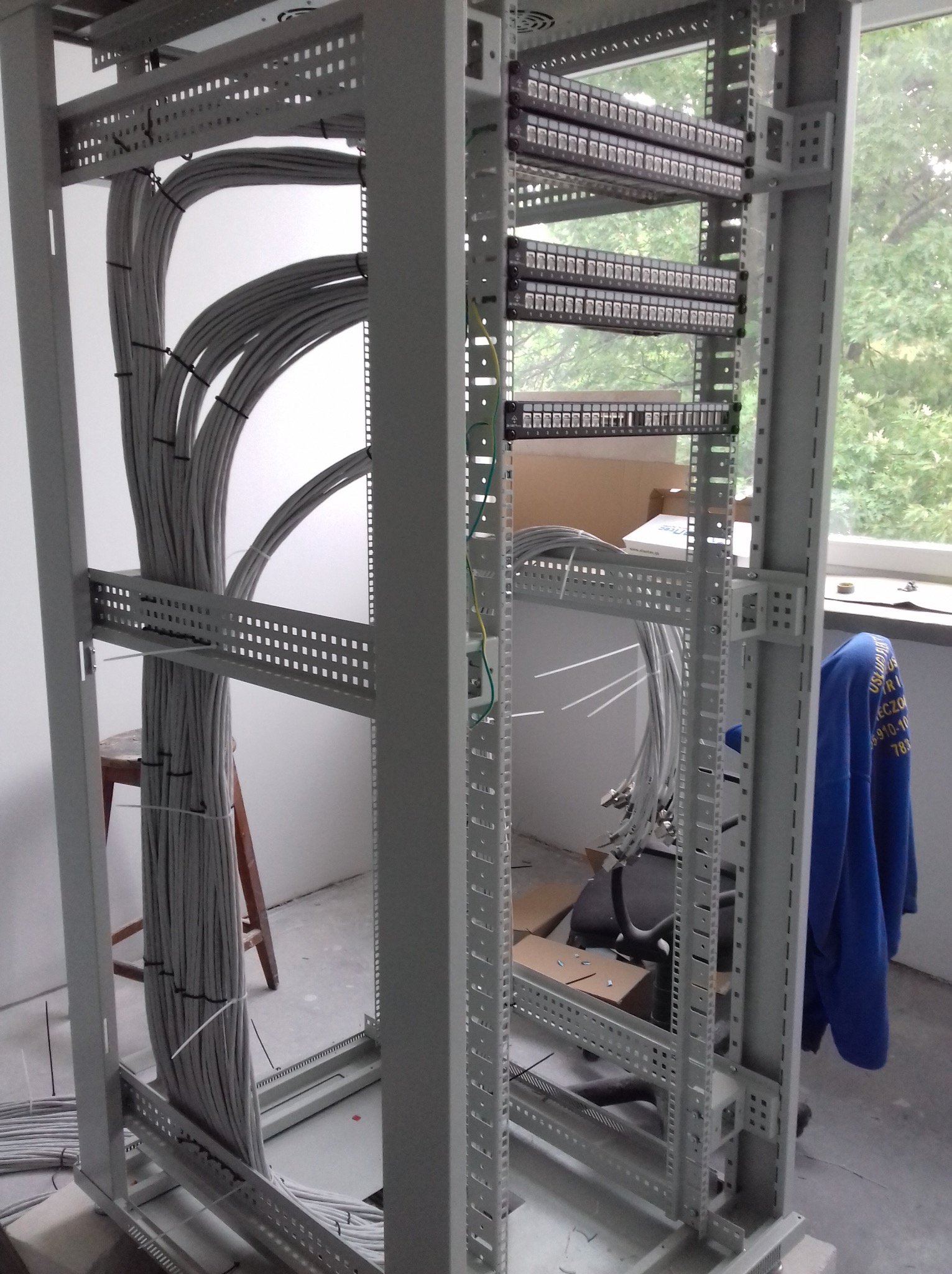 Metalowa szafa rack z uporządkowanymi kablami wewnątrz, widoczne panele krosowe i przewody. W tle okno i fragment pomieszczenia w trakcie prac wykończeniowych.