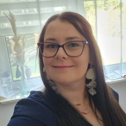 Anna Ciuchta - Uśmiechnięta kobieta w okularach z ciemnymi oprawkami i długimi, jasnymi kolczykami w kształcie liści. Ma długie ciemne włosy, pozuje na tle okna z żaluzjami i wazonem z suszonymi roślinami.