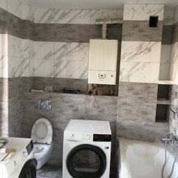 Sylwester Szulga - Wykończona łazienka z marmurowymi płytkami, piecem gazowym, pralką i suszarką. Wanna zabezpieczona folią. Widoczne elementy instalacji.