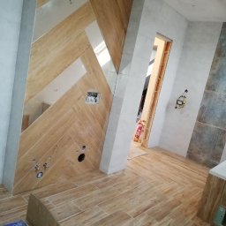 Sylwester Szulga - Ukończona łazienka z drewnopodobnymi płytkami ułożonymi w jodełkę na ścianie i podłodze. Widoczne otwory na armaturę i fragment drzwi.