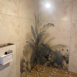 Sylwester Szulga - Układanie płytek z motywem roślinnym w łazience. Widoczne krzyżyki dystansowe na podłodze i fragment instalacji. Beżowe płytki imitujące kamień.