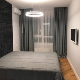 StemBau - Nowoczesna sypialnia w szarościach: łóżko z pikowanym zagłówkiem, okrągła lampa LED, drewniana podłoga w jodełkę i telewizor na ścianie. Minimalistyczny design.