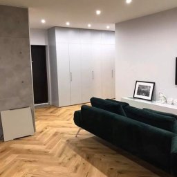 StemBau - Nowoczesny salon z parkietem w jodełkę, ciemnozieloną sofą i minimalistyczną zabudową szaf. Ściana z szarego betonu architektonicznego dodaje surowego charakteru.