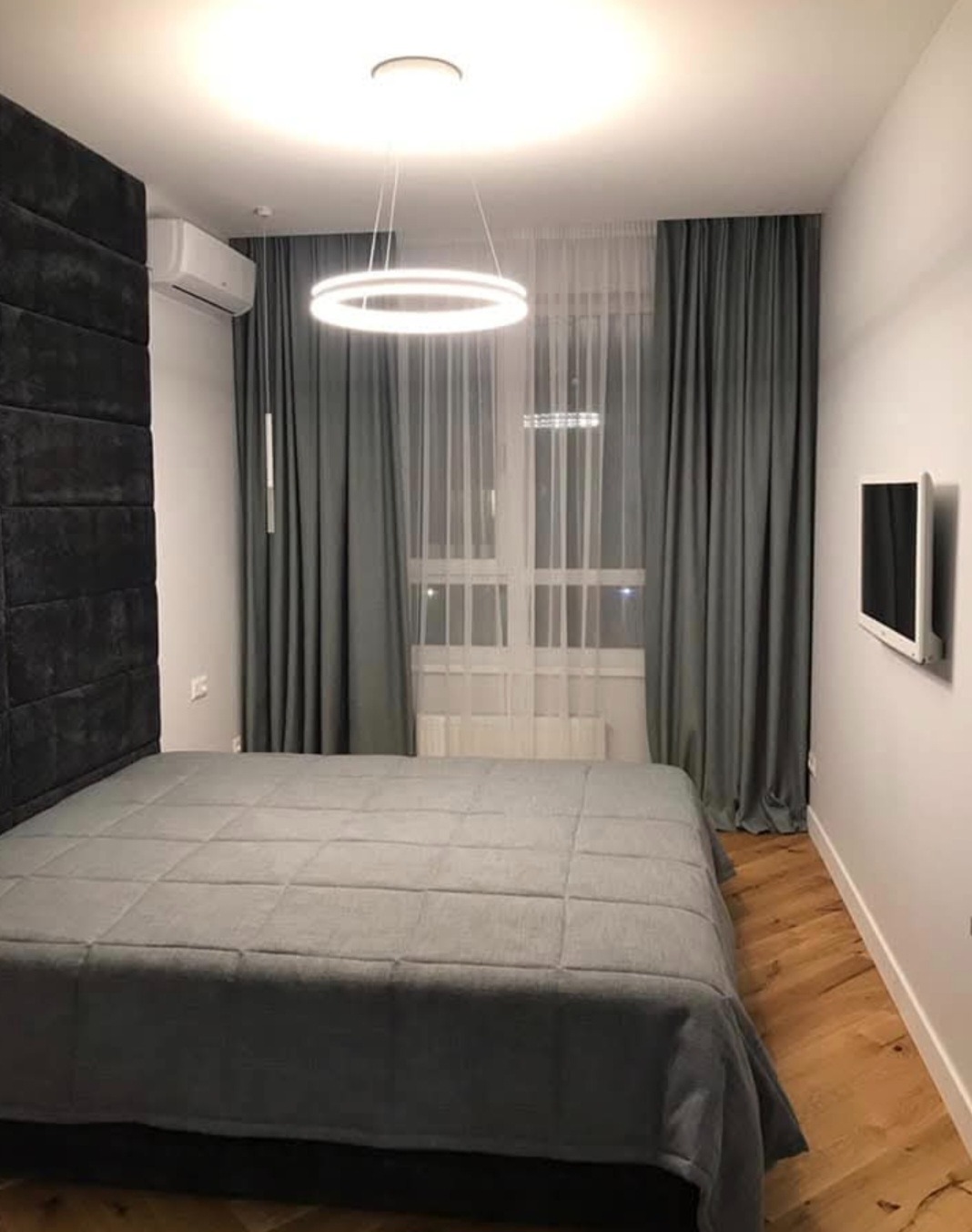 Nowoczesna sypialnia w szarościach: łóżko z pikowanym zagłówkiem, okrągła lampa LED, drewniana podłoga w jodełkę i telewizor na ścianie. Minimalistyczny design.