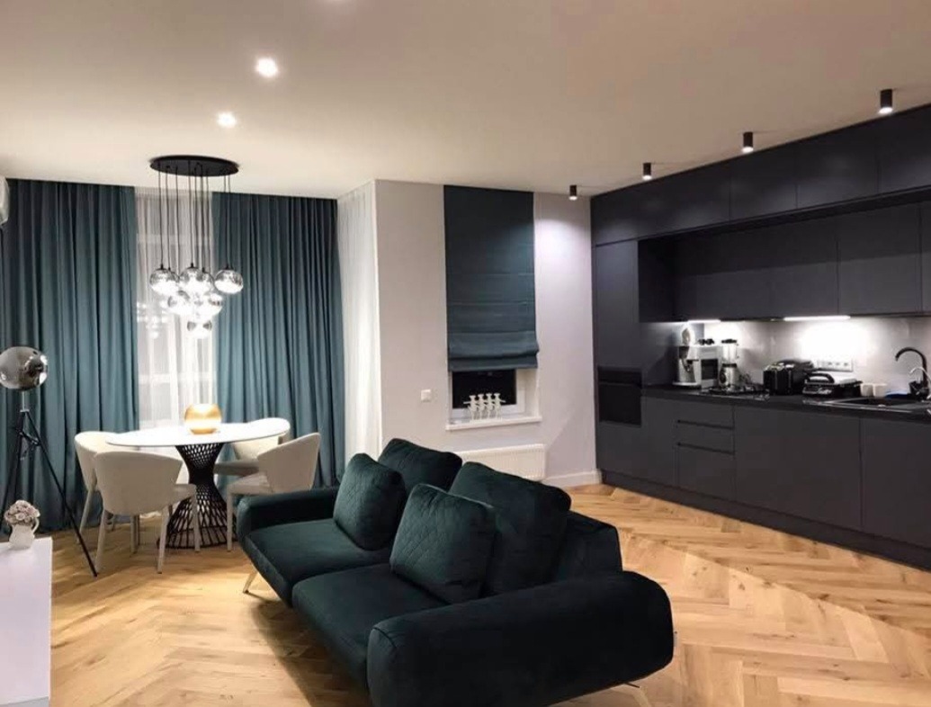 Nowoczesne wnętrze: ciemna zabudowa kuchenna, parkiet w jodełkę, turkusowe zasłony i sofa. Minimalistyczny design z lampami punktowymi i wiszącą nad stołem.