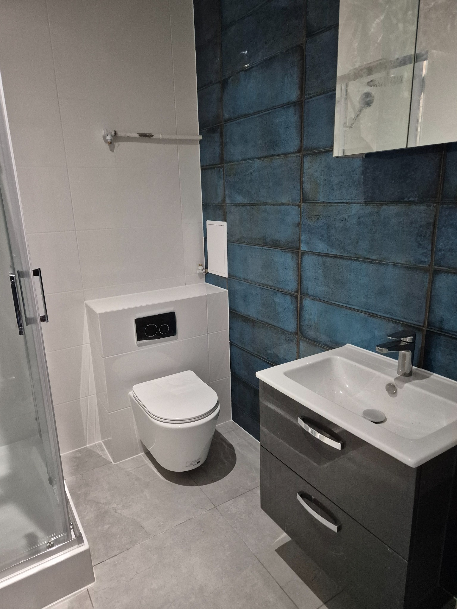 Nowoczesna łazienka po remoncie: biała ceramika, umywalka z szufladami, kabina prysznicowa i ściana z niebieskich płytek imitujących cegłę.