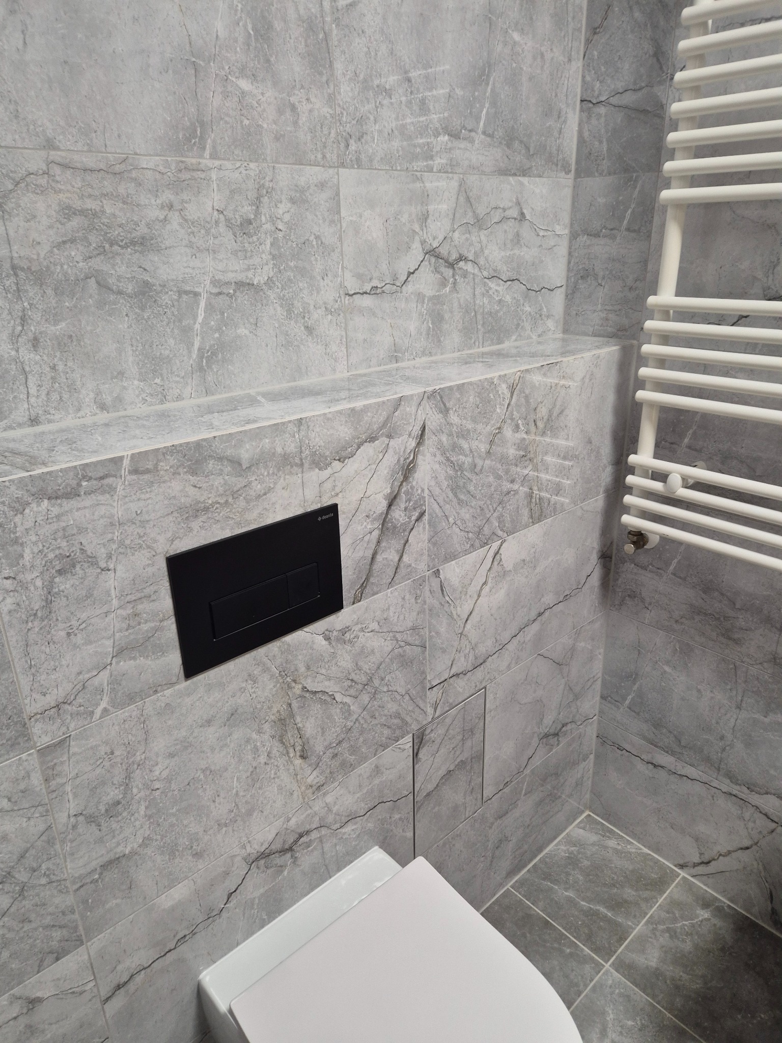 Szare płytki imitujące marmur w łazience z czarnym przyciskiem spłukującym i białym grzejnikiem drabinkowym. Widoczny fragment toalety.