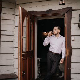 KATARZYNA RYMARCZYK - Elegancki mężczyzna w białej koszuli i wzorzystych spodniach pije drinka, opierając się o futrynę drewnianego domu z numerem 2003. W tle widoczny pies.