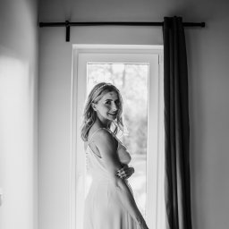 KATARZYNA RYMARCZYK - Czarno-białe zdjęcie uśmiechniętej kobiety w sukience, stojącej w pobliżu okna z zasłoną. Kobieta ma na głowie ozdobę, a na twarzy widoczny jest ślad po makijażu.