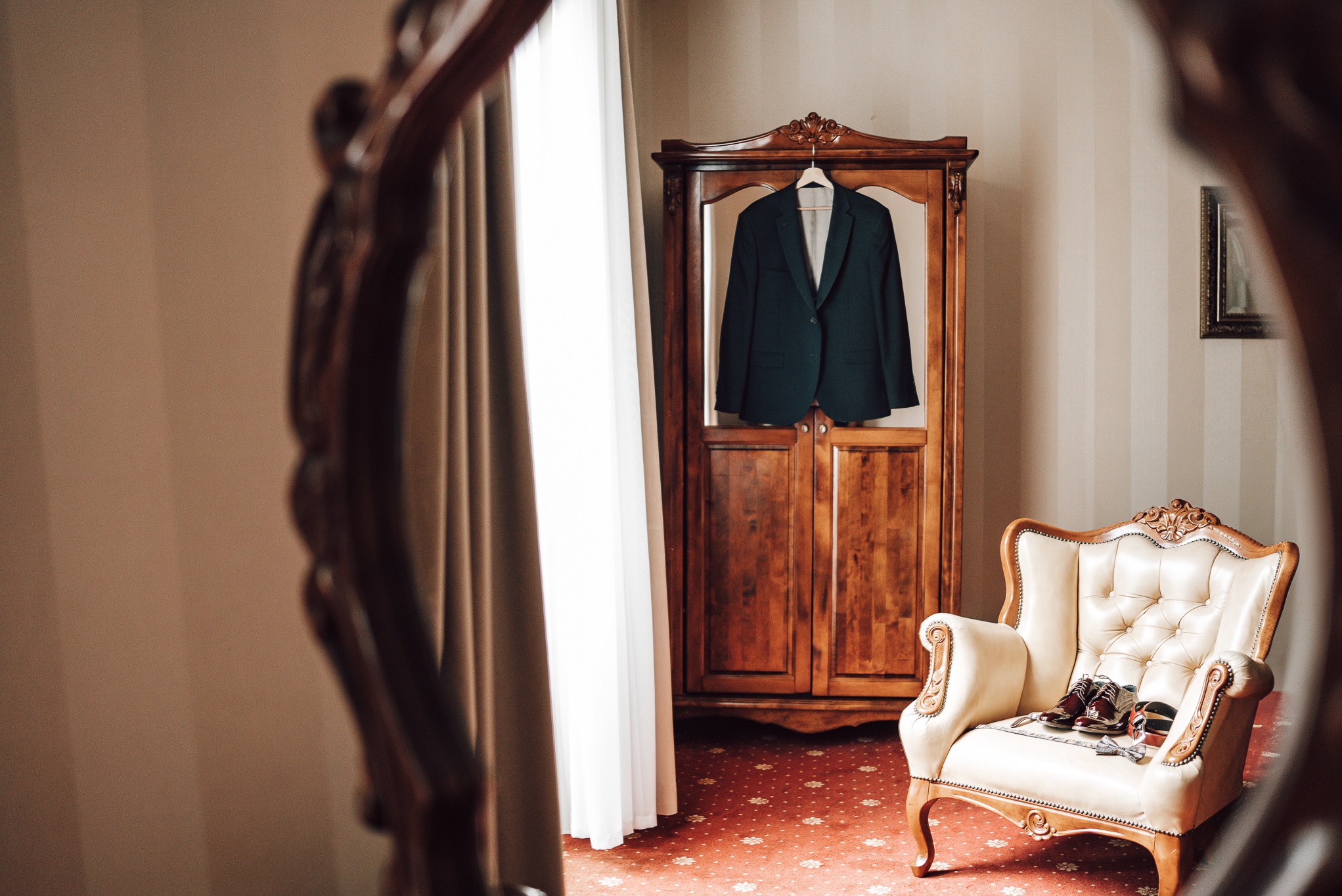 Elegancki pokój widziany w lustrze: garnitur na szafie, kremowy fotel z butami i muszką. Przytulne wnętrze z czerwonym dywanem i subtelnym oświetleniem.