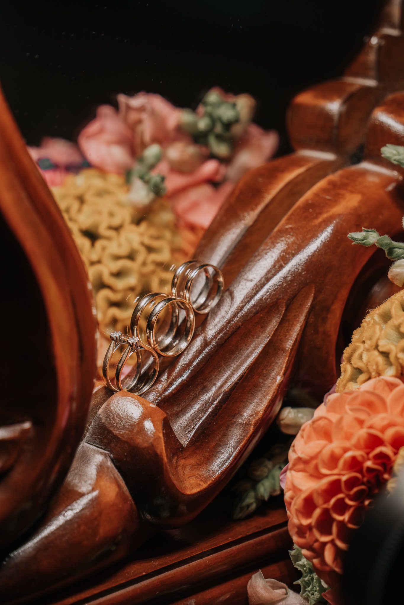 Złote obrączki ślubne z brylantami ułożone na rzeźbionym, drewnianym elemencie dekoracyjnym w otoczeniu kwiatów w odcieniach różu i pomarańczy. Ciepłe, naturalne światło.