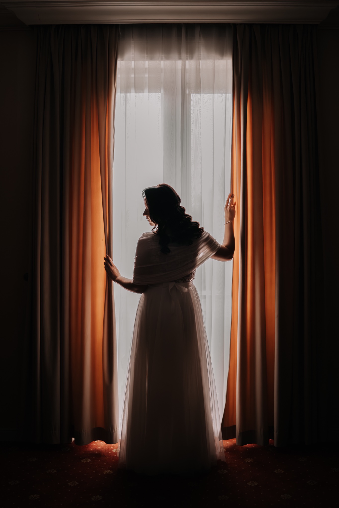 Panna młoda w sukni ślubnej, stojąca tyłem przy oknie z rozchylonymi pomarańczowymi zasłonami, wpadające światło tworzy silny kontrast i podkreśla sylwetkę.