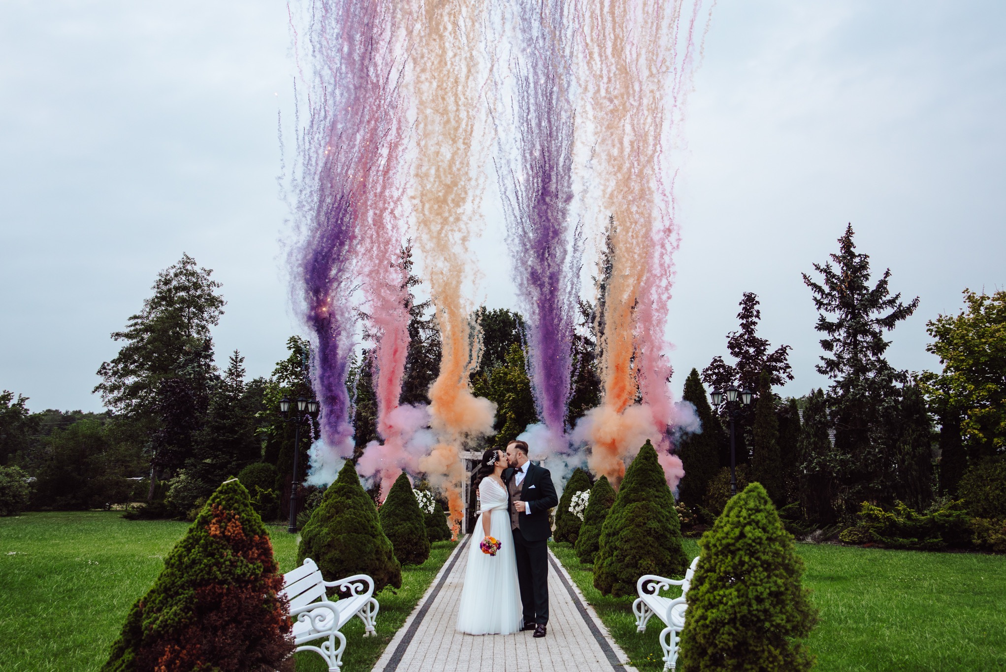 Para młoda całuje się na tle kolorowych fontann dymnych w parku. Panna młoda w białej sukni, pan młody w ciemnym garniturze. Romantyczna sceneria z zielenią i białymi ławkami.