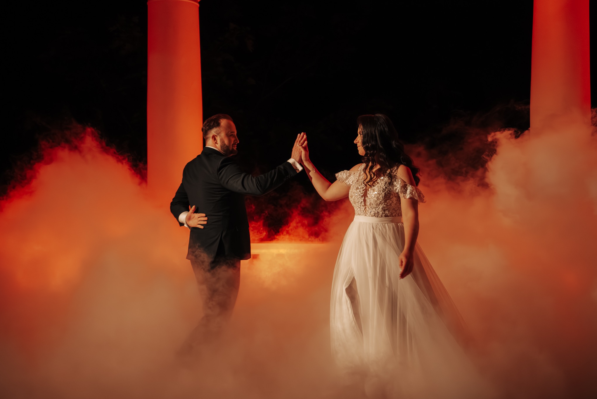 Para młoda w tańcu, otoczona pomarańczową mgłą i kolumnami. Panna młoda w białej sukni, pan młody w czarnym garniturze. Romantyczna atmosfera, ciemne tło.