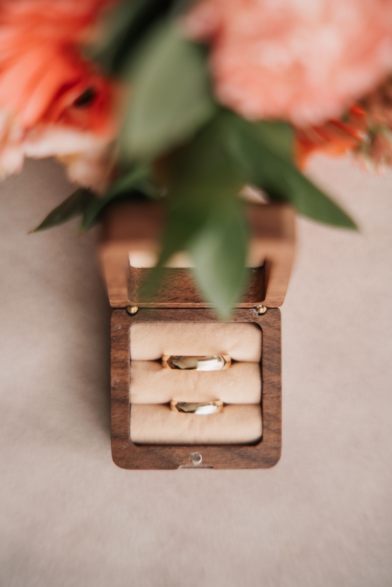 Dwa złote obrączki ślubne w otwartym, drewnianym pudełku z beżowym wnętrzem, na rozmytym tle kwiatów w odcieniach koralowych i zieleni.