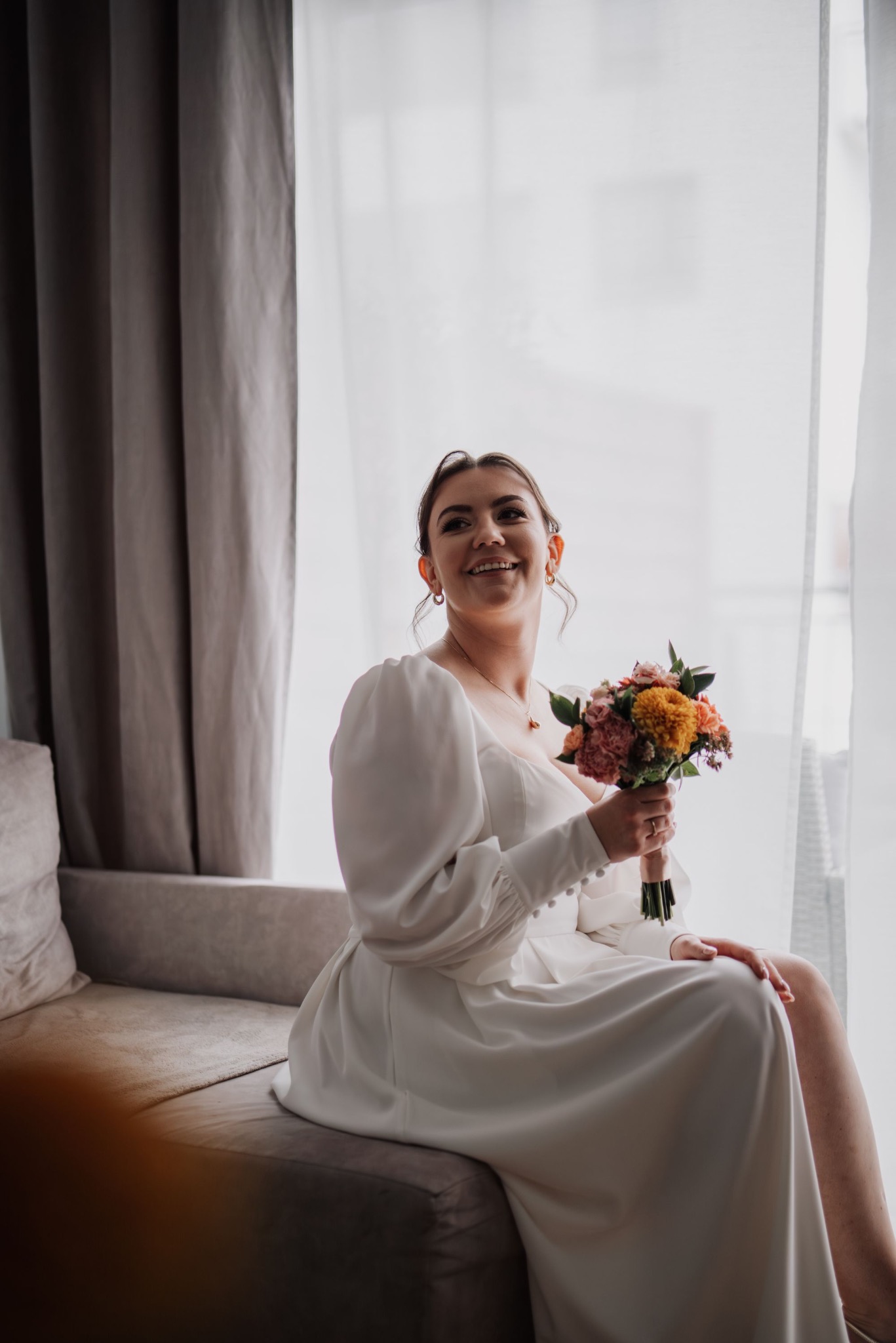 Uśmiechnięta panna młoda w białej sukni z bukietem siedzi na kanapie przy oknie, w tle zasłony. Delikatne światło podkreśla detale sukni i biżuterii.