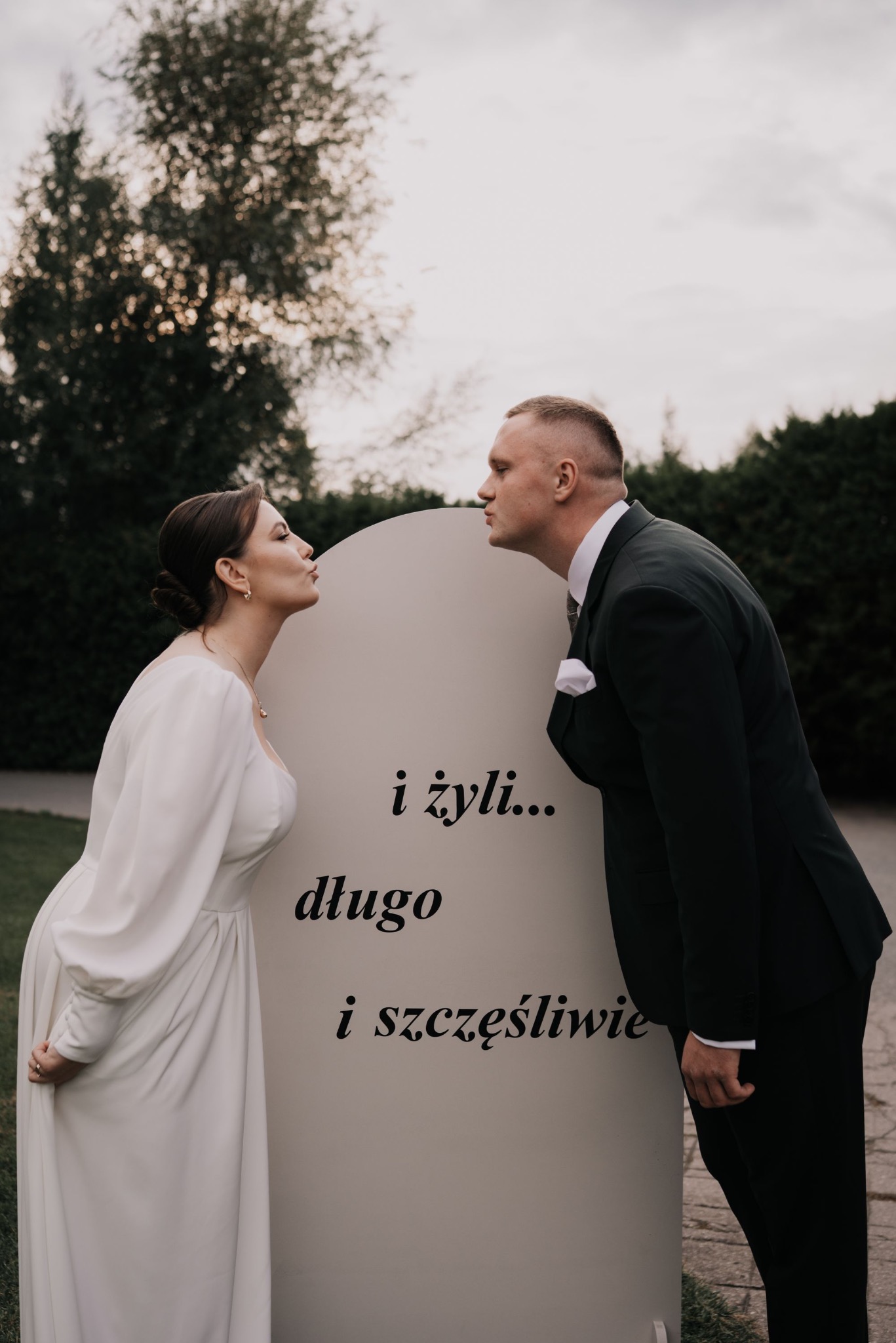 Para młoda w eleganckich strojach, ustawiona po przeciwnych stronach tablicy z napisem 'i żyli długo i szczęśliwie', w plenerze, w delikatnym oświetleniu.