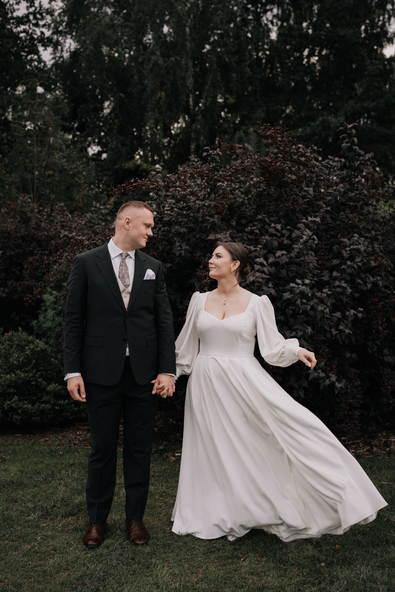 Para młoda trzymająca się za ręce na tle zieleni. Panna młoda w białej sukni z długimi rękawami, pan młody w ciemnym garniturze. Romantyczna atmosfera.