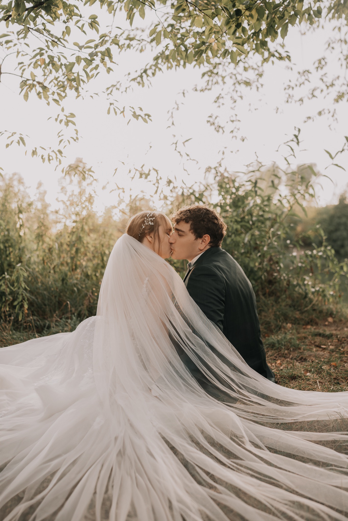 Pocałunek młodej pary siedzącej na ziemi w otoczeniu zieleni. Panna młoda w długiej sukni i welonie, pan młody w ciemnym garniturze. Romantyczna sesja w plenerze.