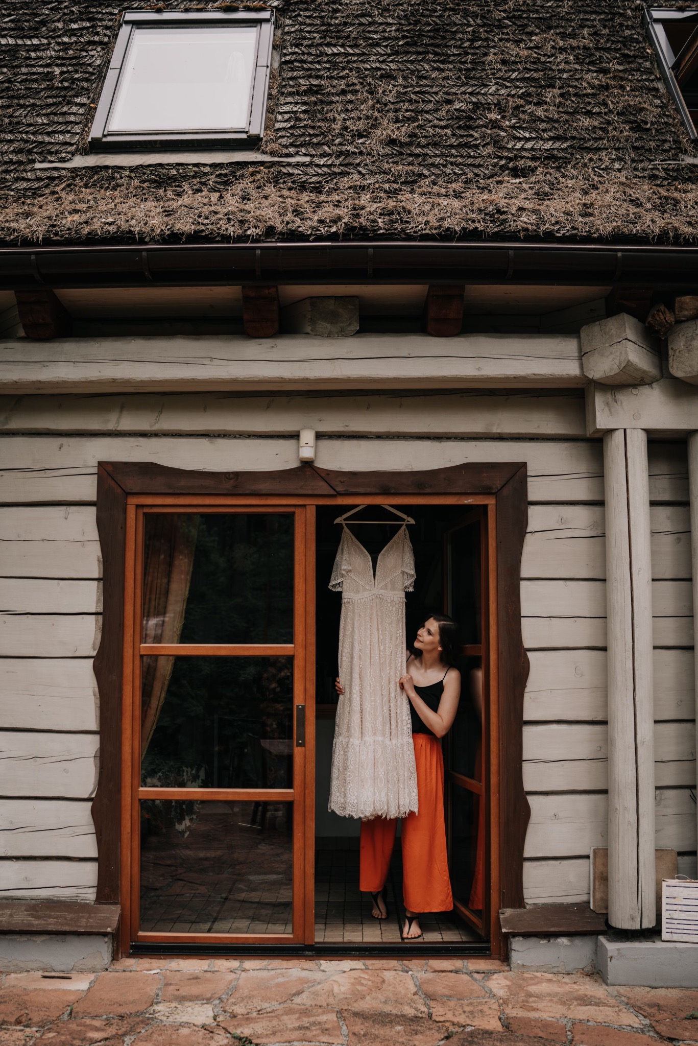 Panna młoda spogląda na suknię ślubną wiszącą w drewnianych drzwiach na tle chaty z gontowym dachem. Romantyczna sceneria, naturalne światło.