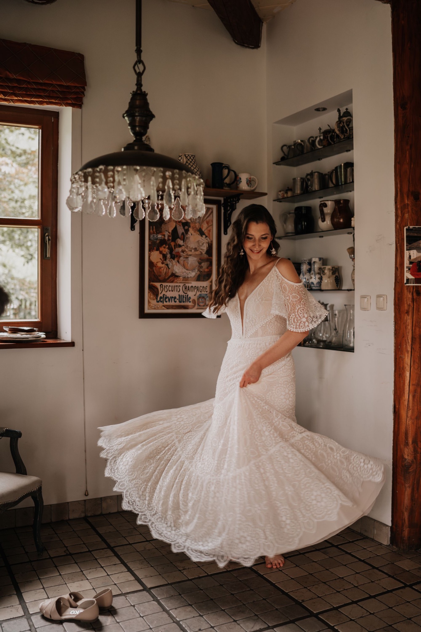Uśmiechnięta panna młoda w koronkowej sukni wiruje w rustykalnym wnętrzu, z widoczną kolekcją naczyń na półkach i butami obok krzesła.