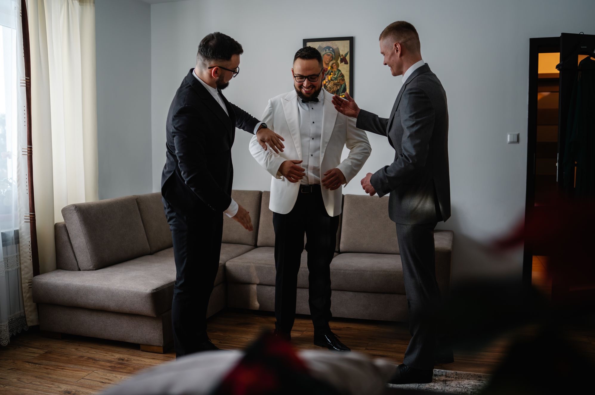 Uśmiechnięty pan młody w białym smokingu, dopinany przez dwóch mężczyzn w garniturach, w jasnym wnętrzu z kanapą i obrazem na ścianie. Przygotowania do ślubu.