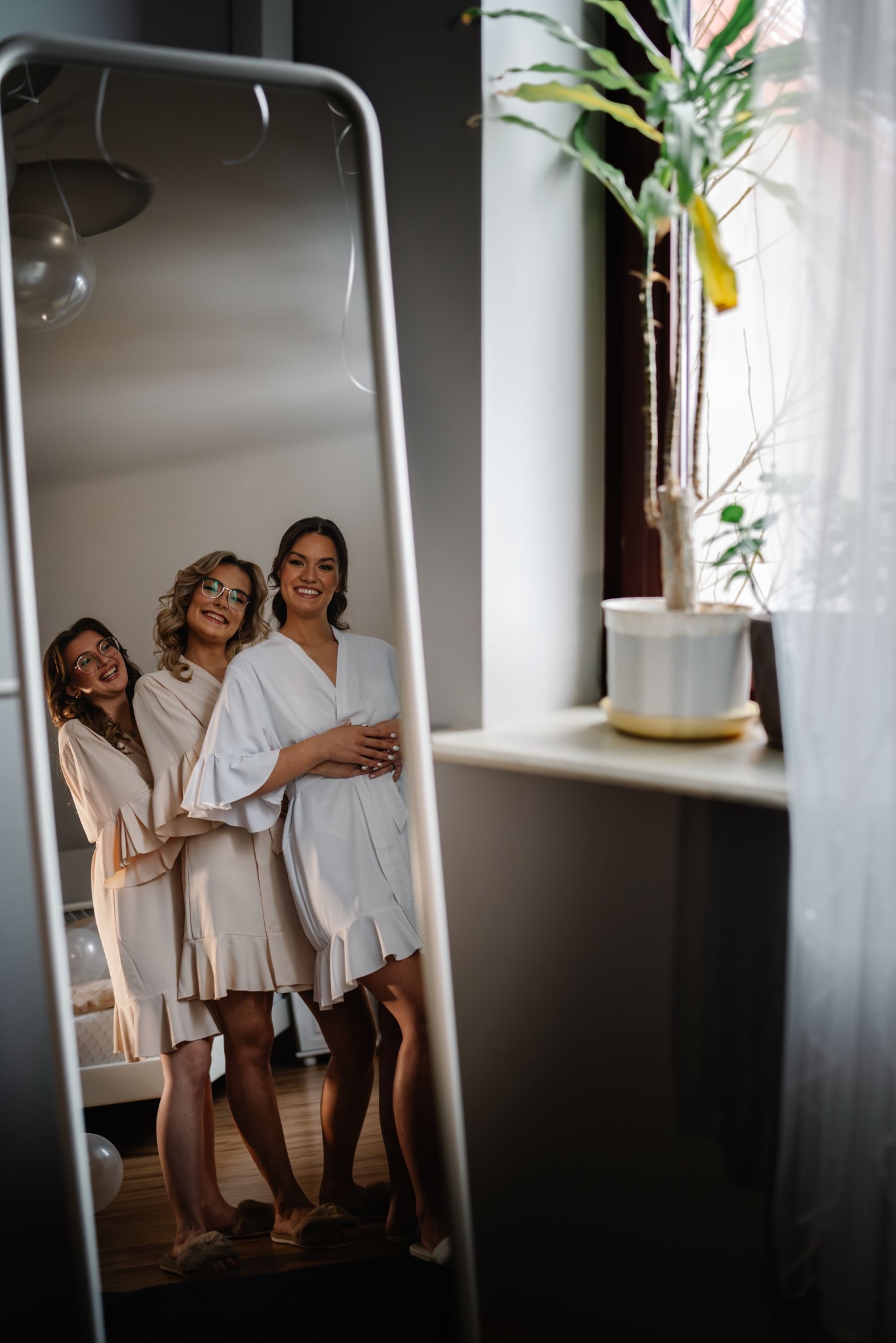 Trzy uśmiechnięte kobiety w szlafrokach, odbite w lustrze. Przygotowania do ślubu, jasne wnętrze z rośliną w tle. Ujęcie z dołu, podkreślające nogi i detale ubioru.