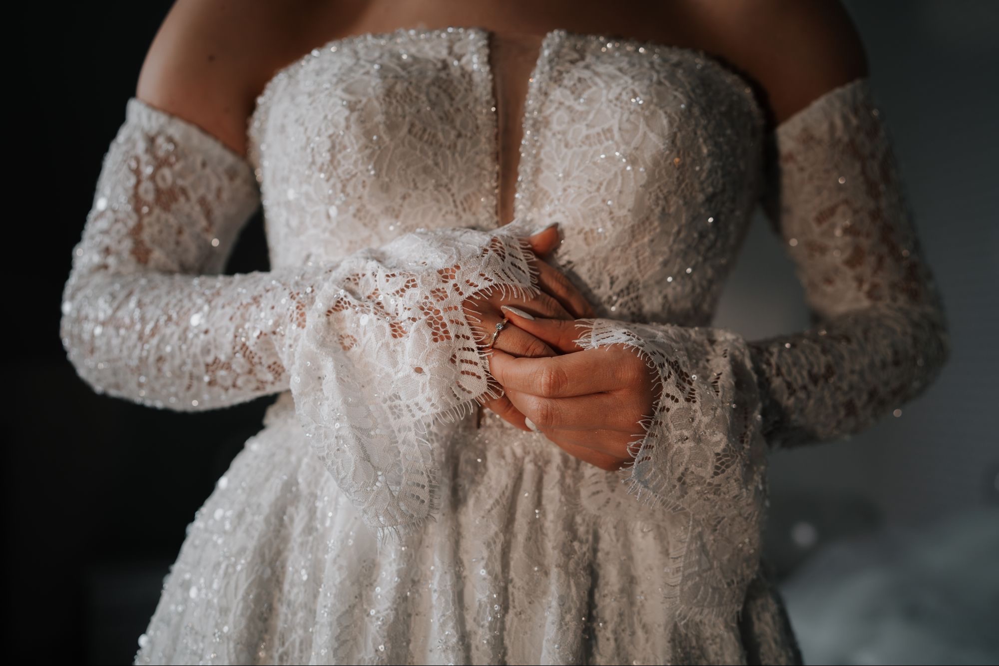 Panna młoda w koronkowej sukni z długimi rękawami, delikatnie dotyka pierścionka zaręczynowego. Subtelne światło podkreśla detale sukni i dłoni.
