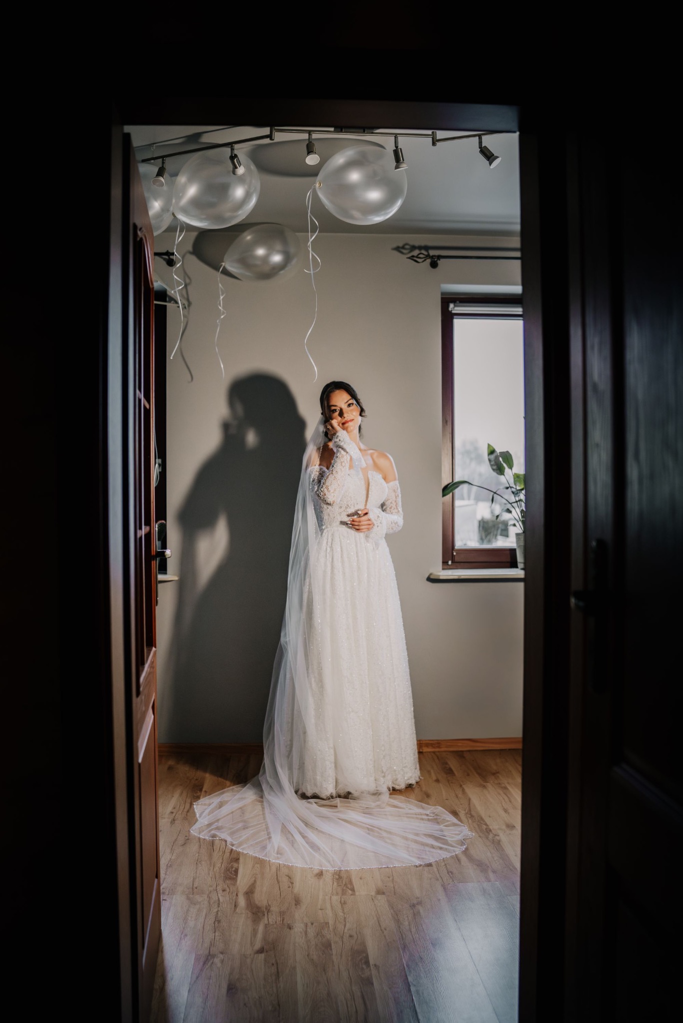 Panna młoda w białej sukni pozuje w pokoju z balonami, widziana zza otwartych drzwi. Delikatne światło pada na suknię i twarz, tworząc intymną atmosferę.