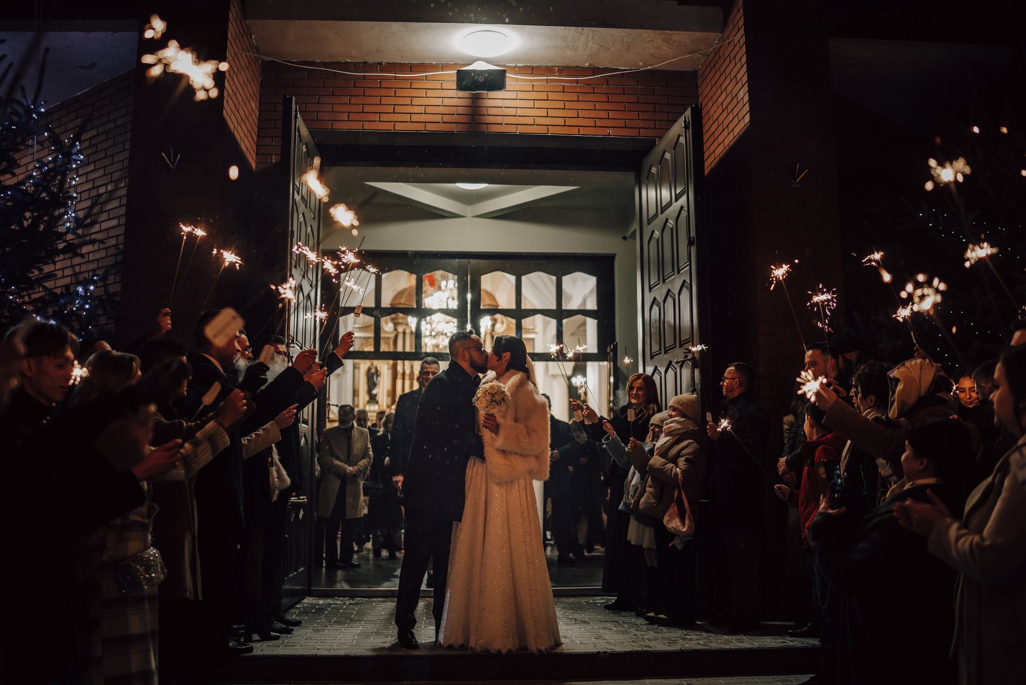 Pocałunek nowożeńców na tle kościoła, otoczonych gośćmi z zimnymi ogniami. Wieczorowa atmosfera, ciepłe światło, dekoracje świąteczne w tle.