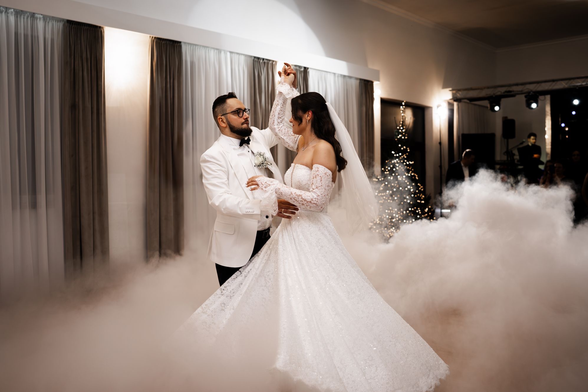 Para młoda w białych strojach tańczy pierwszy taniec w dymie na tle dekoracji ślubnej z choinką i zasłonami. Pan młody w okularach.