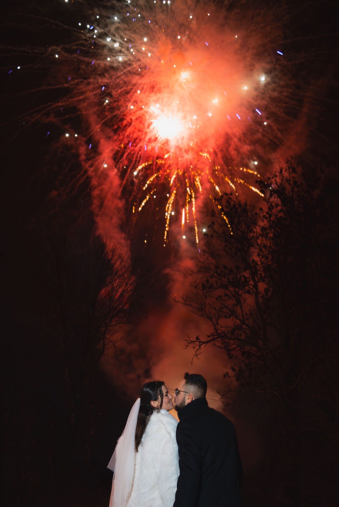 Pocałunek pary młodej na tle nocnego nieba rozświetlonego czerwonymi fajerwerkami. Panna młoda w białym futerku i welonie, pan młody w czarnym płaszczu.