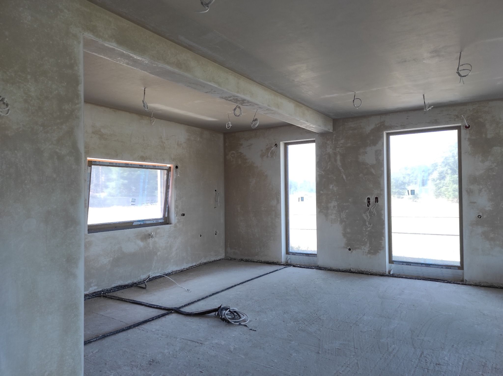 Wnętrze po tynkowaniu maszynowym: surowe ściany, okna z widokiem na zieleń, kable na podłodze. Widoczne niedoskonałości faktury tynku.