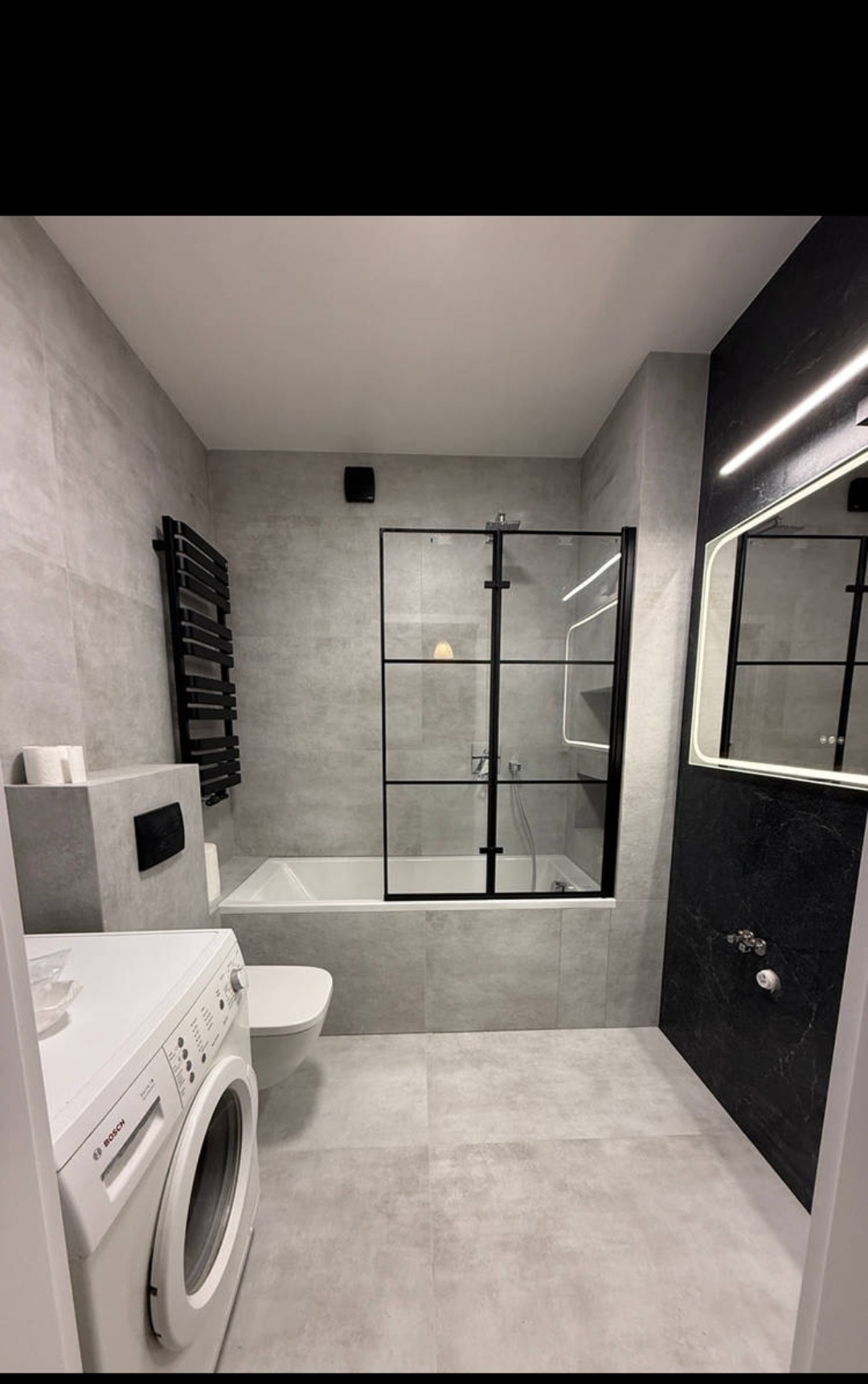 Nowoczesna łazienka w szarościach z czarnymi akcentami: kabina prysznicowa typu walk-in, grzejnik drabinkowy i pralka Bosch. Minimalistyczny design.