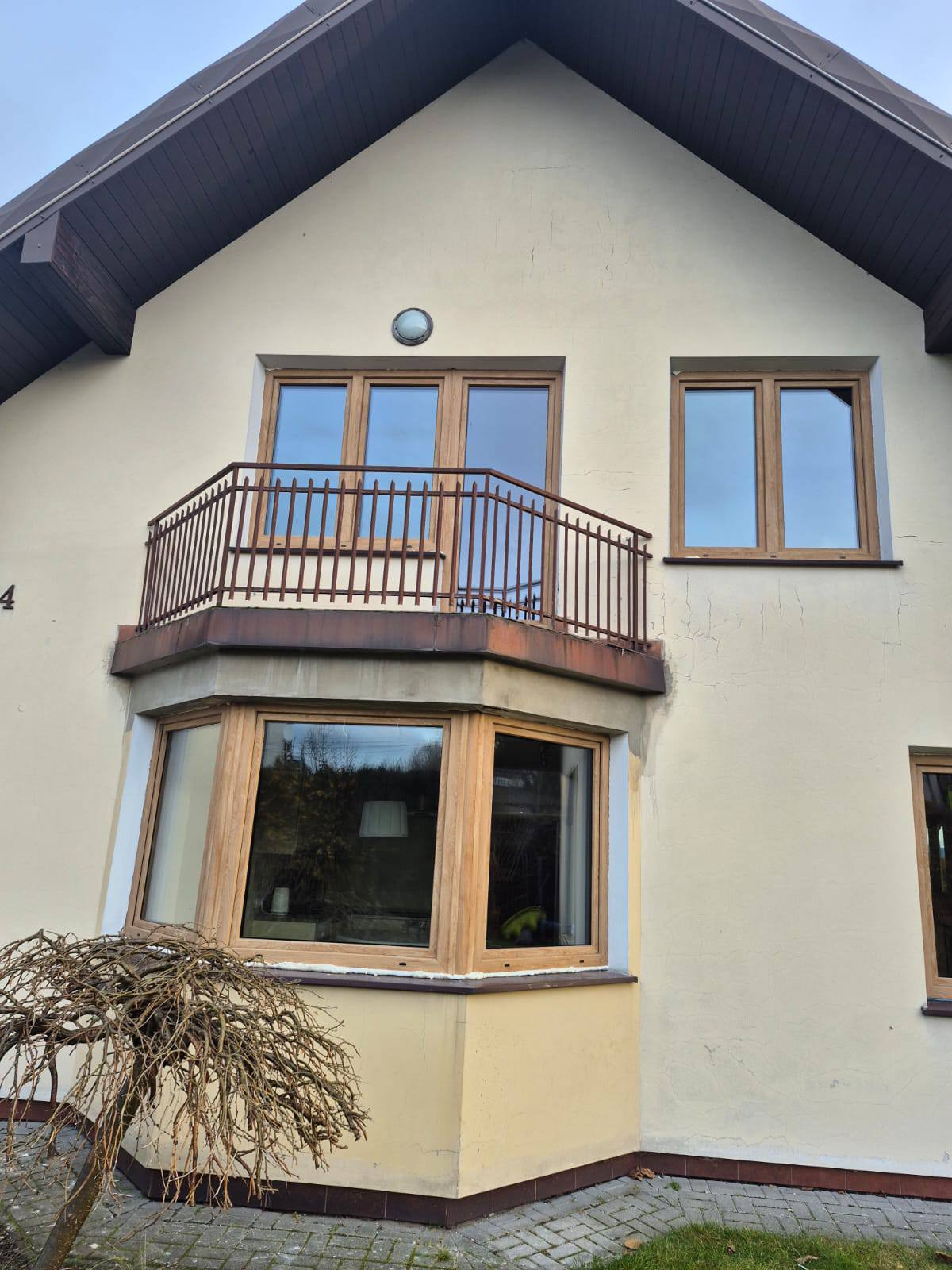 Elewacja domu z oknami PCV w drewnianej okleinie i balkonem z metalową balustradą. Widoczne pęknięcia na elewacji i drzewko na pierwszym planie.