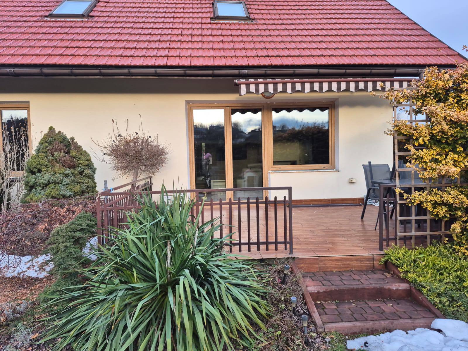 Dom z beżową elewacją, drewnianym tarasem i oknami. Widoczny markiza nad oknem tarasowym, roślinność w ogrodzie i fragment czerwonego dachu z oknami połaciowymi.